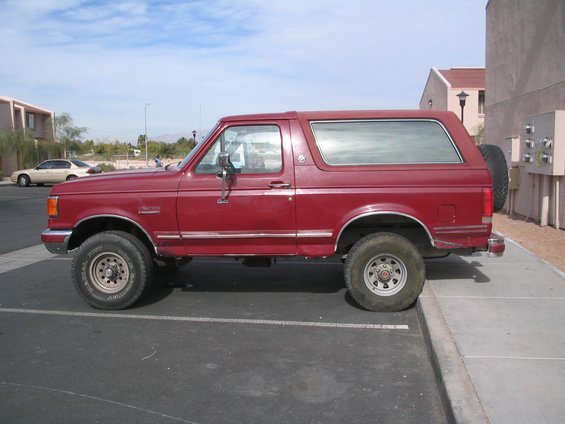 Side view of a red 1991 Ford Bronco parked in a lot. It has a spare tire mounted on the back
