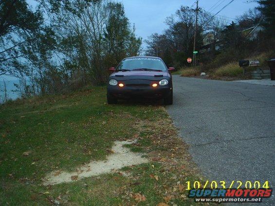 Front view of a dark-colored car with a black car bra and yellow parking lights, parked on a grassy area next to a road