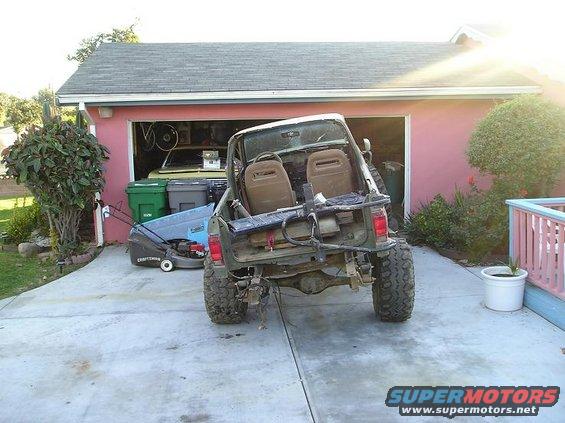 Off-road SUV with lifted suspension and large tires parked in front of a garage. It has a roll bar and two bucket seats visible