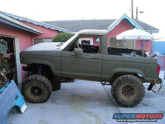 Olive green lifted truck with large tires, missing front bumper and doors, parked in driveway