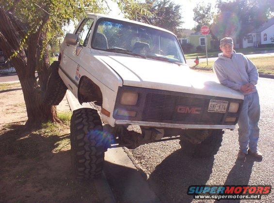 guy leaning on front of white GMC pickup truck with front tire up in a tree