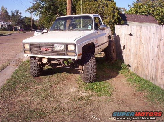 Lifted 1985 GMC pickup truck parked on grass next to a wooden fence. License plate reads RMF K74