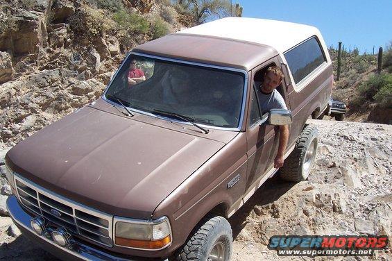 Brown Ford Bronco with a white top driving on a rocky trail, driver leans out window