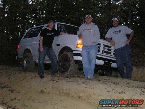 Three guys stand around a white Ford Bronco on a dirt path in the woods