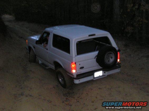 White Ford Bronco driving on a dirt road at dusk. Spare tire mounted on the back