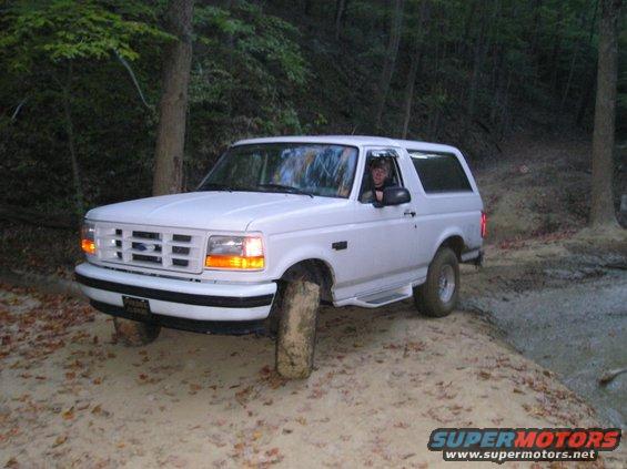 White Ford Bronco driving on a dirt trail with trees around. Driver is visible