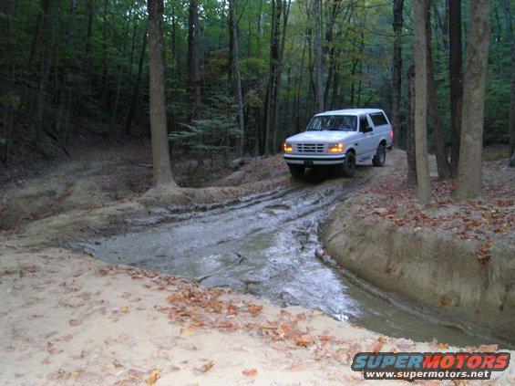 White 1996 Ford Bronco driving through a muddy forest trail