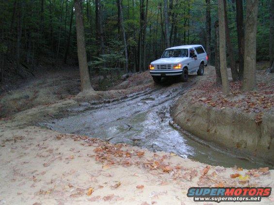 White 1996 Ford Bronco driving through mud on a wooded trail