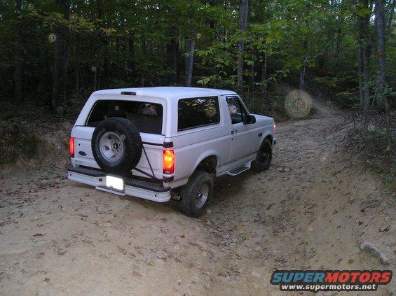 1996 Ford Bronco driving on a dirt trail through a forest, spare tire mounted on the rear door
