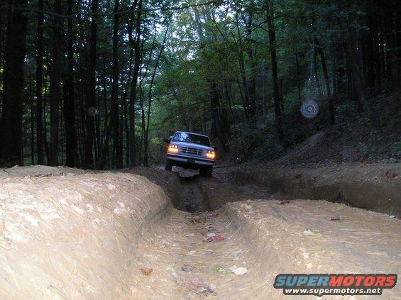 Ford Bronco driving through a muddy rutted trail in a wooded area. Headlights are on