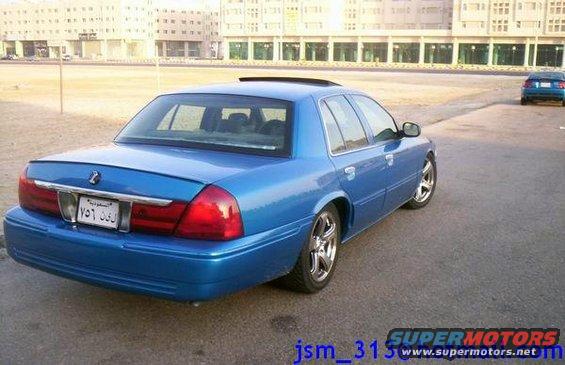 Blue sedan with a spoiler and aftermarket wheels parked on asphalt, with a second blue car in the background