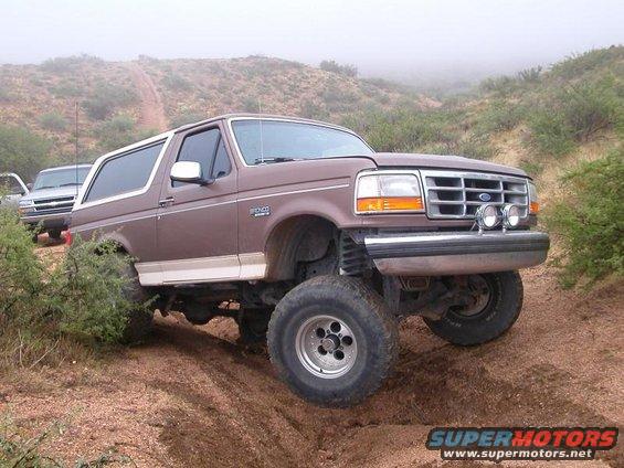 Brown Ford Bronco with lifted suspension driving on a dirt trail with another vehicle behind it