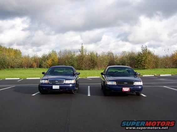 Two blue Ford Crown Victorias parked in a lot, front view. One has a white plate, the other a red plate