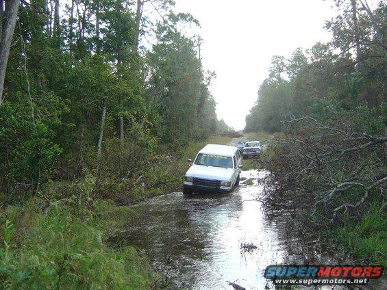 White 1993 Ford Bronco driving through muddy road with trees and fallen branches