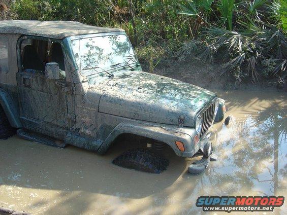 Mud-covered Jeep Wrangler stuck in a muddy ditch with vegetation in the background. Supermotors.net logo visible