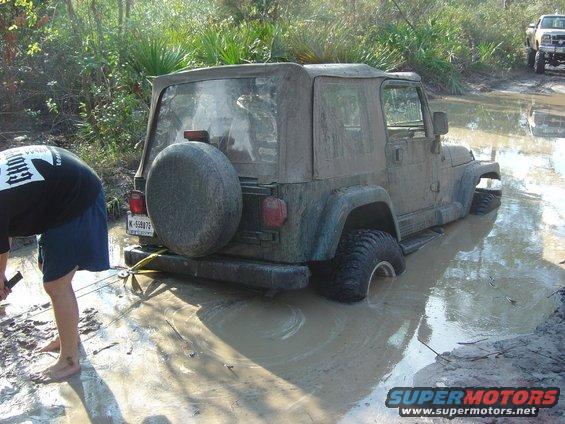 Muddy Jeep Wrangler stuck in water with a person trying to pull it out. Another truck is visible in the background