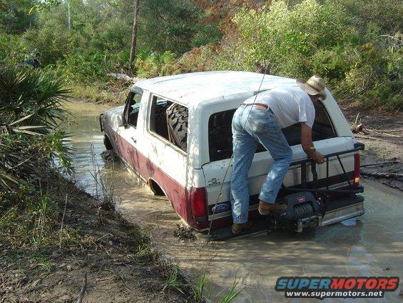 Man climbing onto a white and maroon Ford Bronco stuck in a muddy ditch. Spare tire visible inside