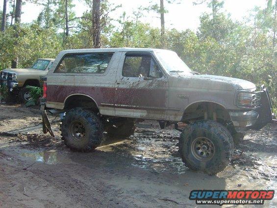 Mud-covered 1992 Ford Bronco with lifted suspension and off-road tires, parked in a muddy wooded area next to another truck