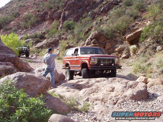 Red Ford Bronco driving on rocky terrain, man standing nearby