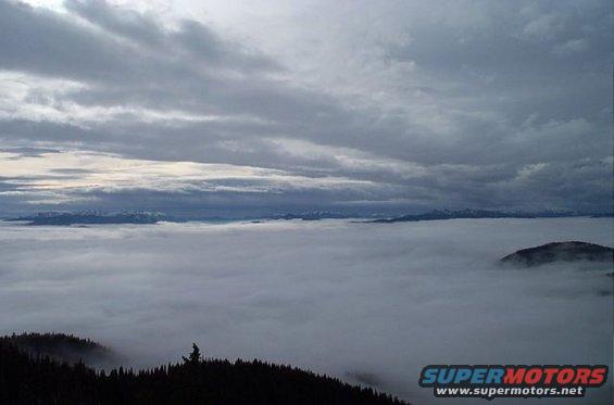 Overcast sky above a thick layer of clouds with mountains visible in the distance. A dark, forested hill is in the foreground