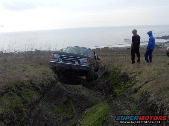 Ford pickup truck driving down a muddy ditch near the ocean. Two people watch