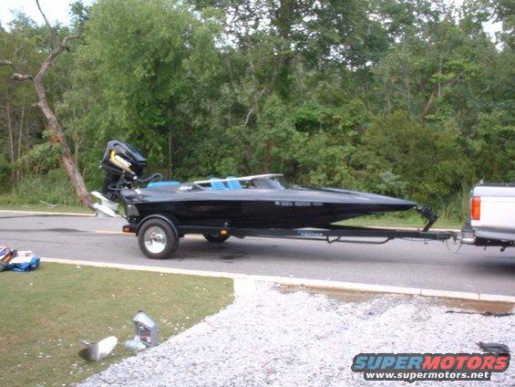 Black speed boat on a trailer, towed by a white pickup truck. A spare propeller and oil bottles are on the grass