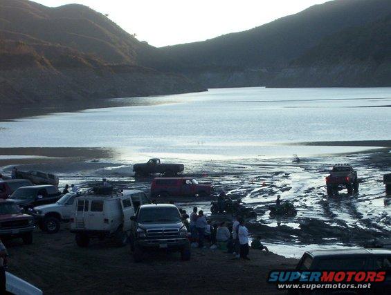 Several off-road vehicles parked on muddy ground near a large body of water with hills in the background