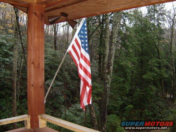 American flag hanging from a pole on a wooden porch, with a forest in the background. Supermotors.net logo visible