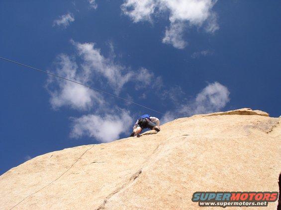 Rock climber ascending a large, tan rock face under a blue sky with white clouds. A rope is visible