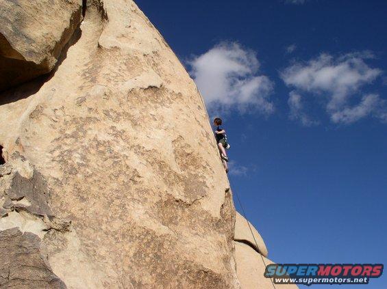Rock climber ascends a large boulder under a blue sky with scattered clouds. Watermark reads SUPERMOTORS.NET