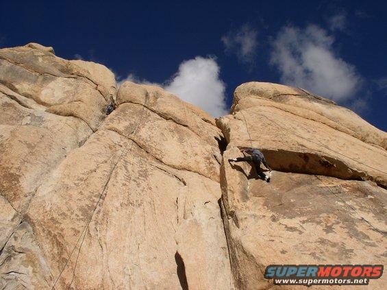Two people climbing a large rock formation under a blue sky with clouds. A watermark reads SUPERMOTORS.NET