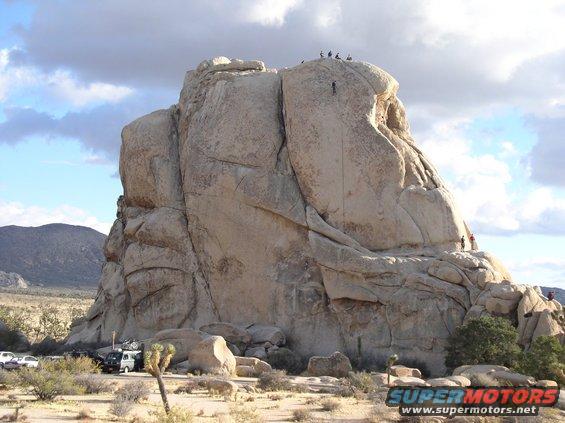 Desert landscape with large rock formation, climbers on the rock, and several vehicles parked at the base