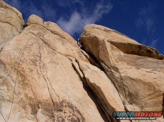 Rock climbers ascending a large rock formation under a blue sky with clouds. Watermark reads SUPERMOTORS.NET