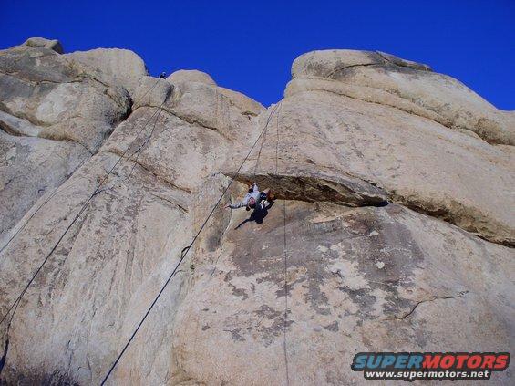 Rock climbers ascending a large rock formation under a clear blue sky. A watermark reads SUPERMOTORS