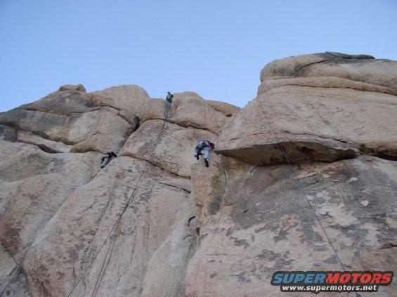 Rock climbers ascend a large boulder formation under a clear blue sky; a watermark reads SUPERMOTORS.NET