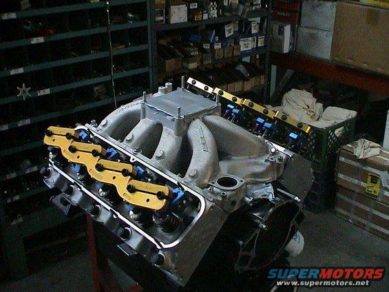 V8 engine block with intake manifold, yellow rocker arms, and blue fuel injectors. Background shows shelves with parts