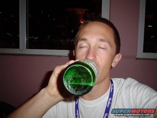 Man drinking from a green bottle, eyes closed, wearing a white t-shirt with a blue lanyard. Text 'SUPERMOTORS' visible
