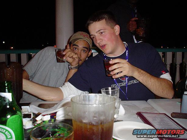 Two guys are sitting at a table with drinks. One is wearing a tan baseball cap and the other a navy blue t-shirt