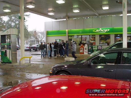 Dark sedan in foreground, red car hood in foreground, gas pumps, and people standing under a canopy at a shop