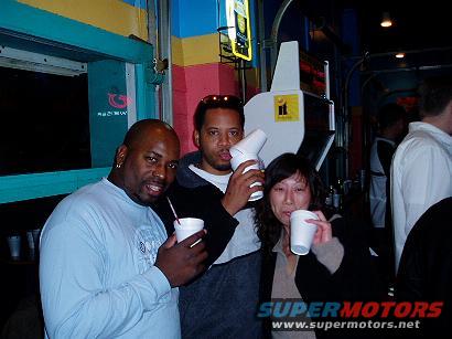 Three people holding drinks indoors, with a "SUPERMOTORS" logo visible