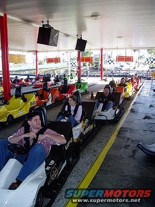 Go-karts lined up on a track, with people riding in them. The go-karts are colorful and varied in design