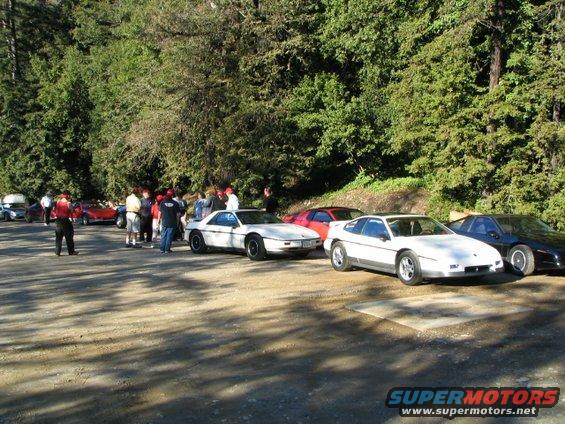Three white Pontiac Fieros and one black Pontiac Fiero parked on a dirt road with trees in the background