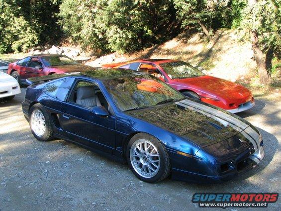 Blue Pontiac Fiero with silver racing stripes and aftermarket wheels, parked on gravel