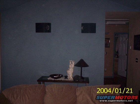 Room interior with couch, table with lamp and statue, and speakers on wall. Doorway visible in background
