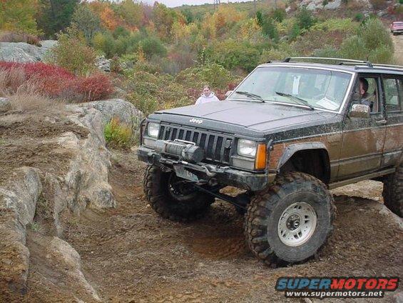 Jeep Cherokee XJ with large tires off-roading on a dirt trail with rocks and fall foliage