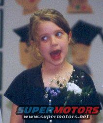 Young girl wearing a black dress and graduation cap, looking to the side with mouth open. Watermark 'SUPERMOTORS www.supermotors.net' at bottom