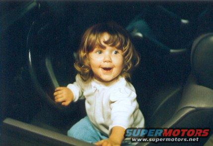 Child sitting in driver's seat, holding steering wheel. "SUPERMOTORS" logo visible