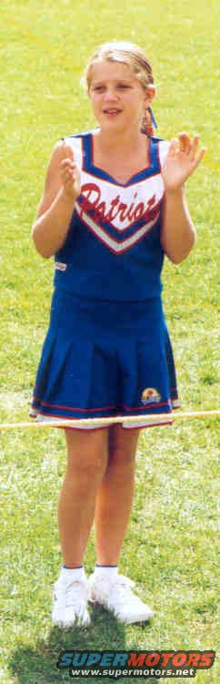 Young girl in a blue and white 'Patriots' cheerleader uniform stands on grass, clapping
