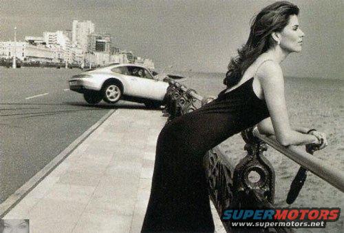 Woman in black dress leans on railing, looking out at sea. Silver Porsche drives on road behind her. "SUPERMOTORS www.supermotors.net" logo visible