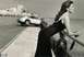 1990 Ford Bronco - A woman observes a Porsche 911 as it drives past a coastal city.